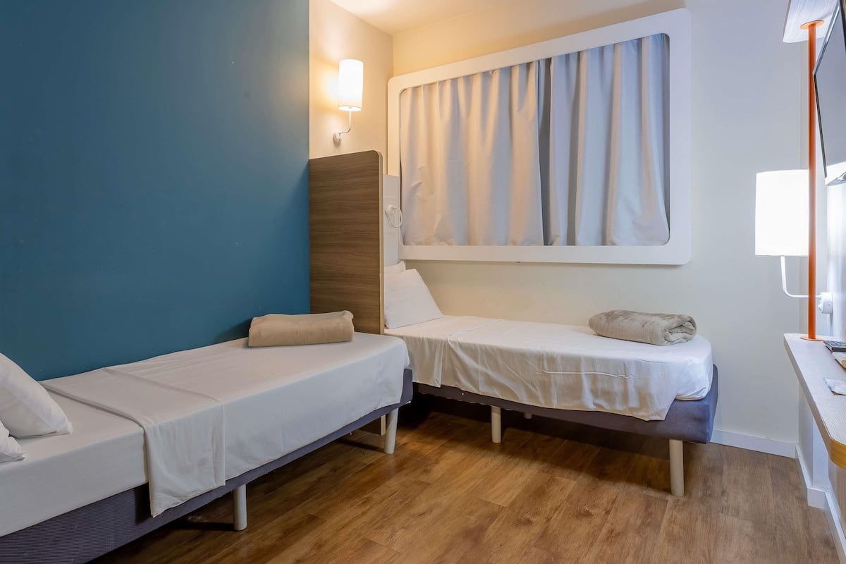 A small, compact room with two single beds, a wooden floor, a window with curtains, and a desk with a lamp.