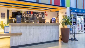 A hotel reception desk with a 'Priority Welcome' sign, a large potted plant, and a vending machine. - ibis budget Patos de Minas (Patos de Minas)