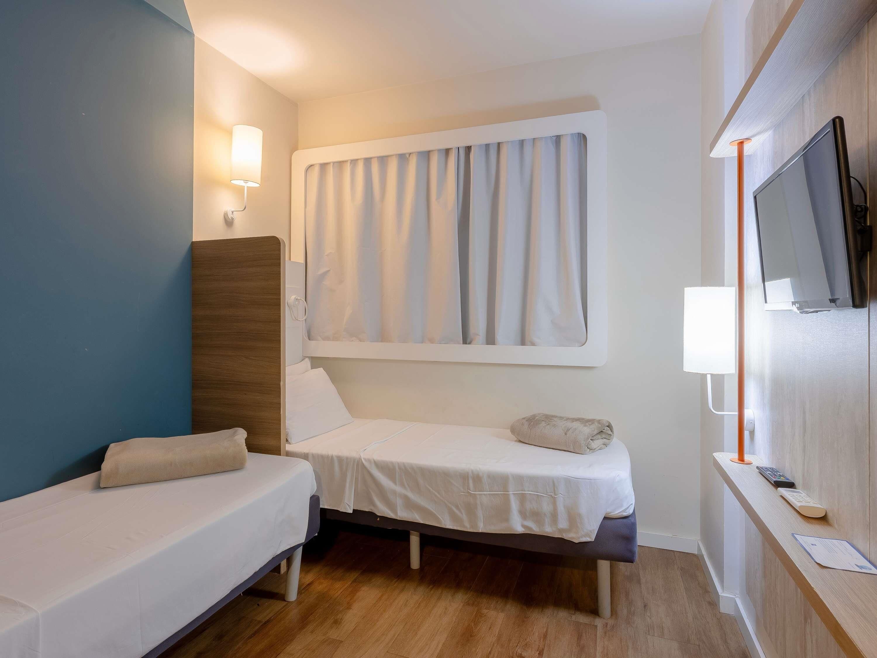 Et lille hotelværelse med to senge, et vindue med gardiner, et tv ophængt på væggen og et natbord med lampe.