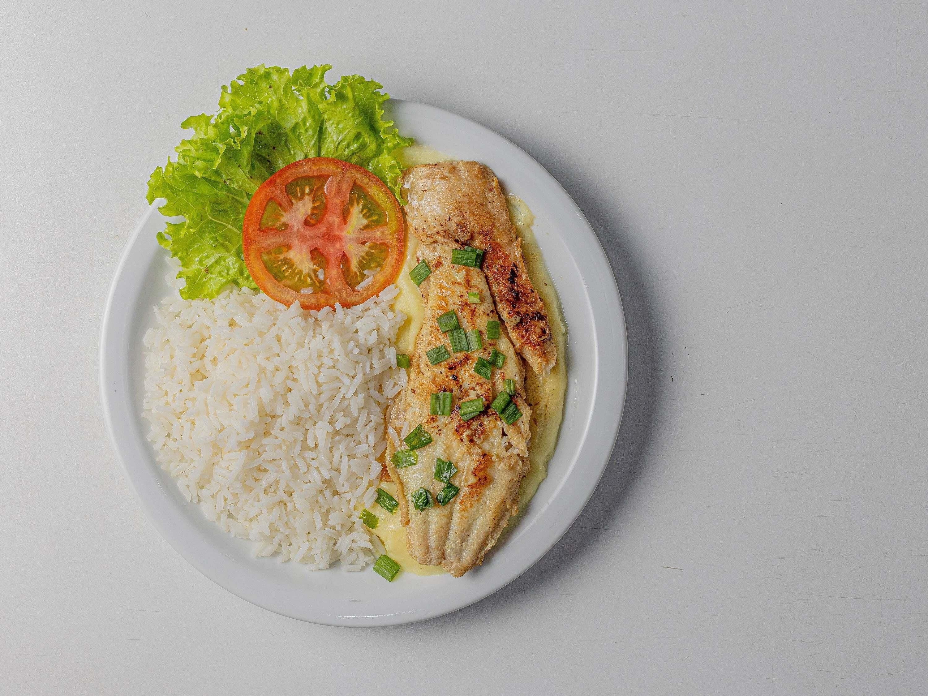 A plate with white rice, a piece of grilled fish, a slice of tomato, and a leaf of lettuce.