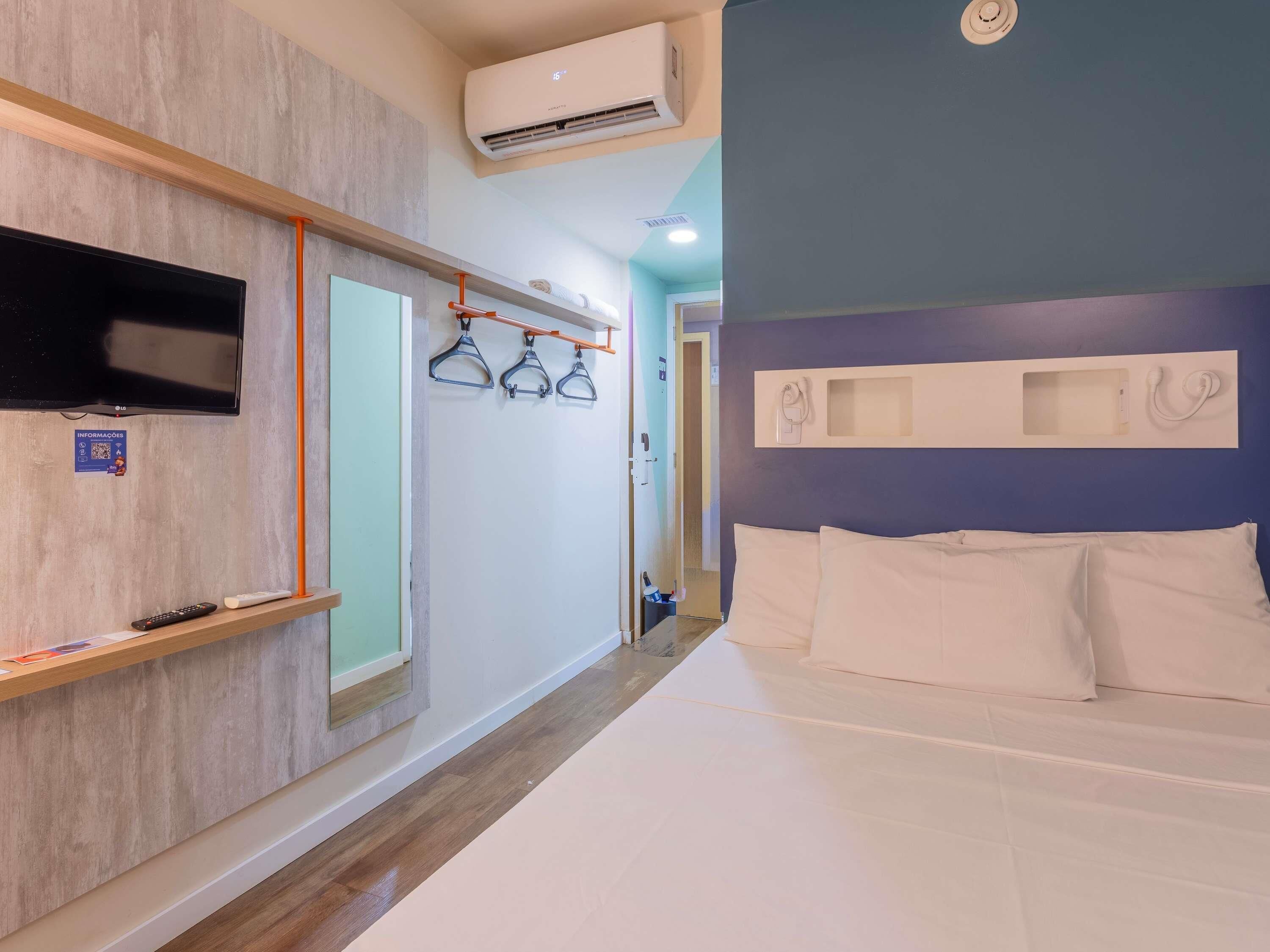 A hotel room with a bed, a TV, a wall-mounted air conditioner, a coat rack, and a door.