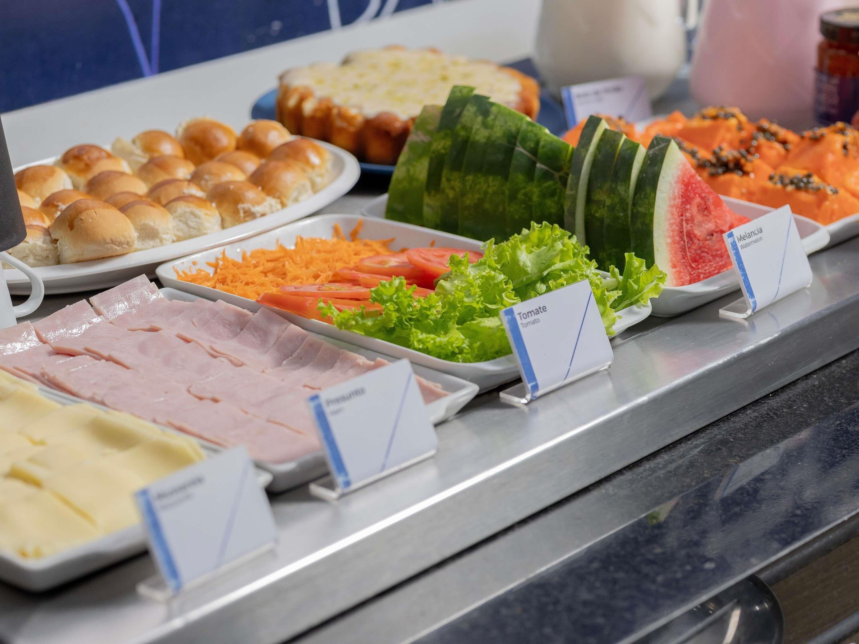 A buffet with various food items including ham, cheese, bread, lettuce, tomatoes, watermelon, and a dish labeled 'Melanca Watermelon'.