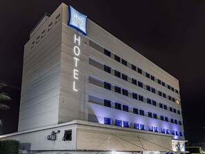 Um hotel com vários andares, com uma placa grande que diz 'HOTEL' na fachada, uma placa 'ibis budget' acima e um design arquitetônico moderno com linhas horizontais e janelas iluminadas.