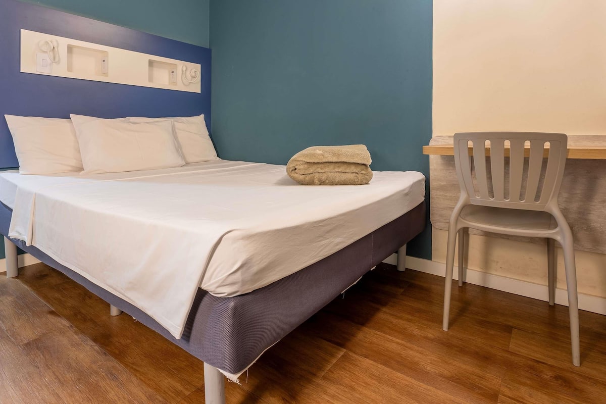 A bed with white bedding, a folded towel, a grey chair, and a wooden floor.