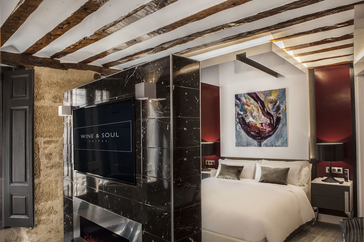 A modern hotel room with a large black marble fireplace, a bed with white linens, a painting on the wall, and wooden beams on the ceiling.