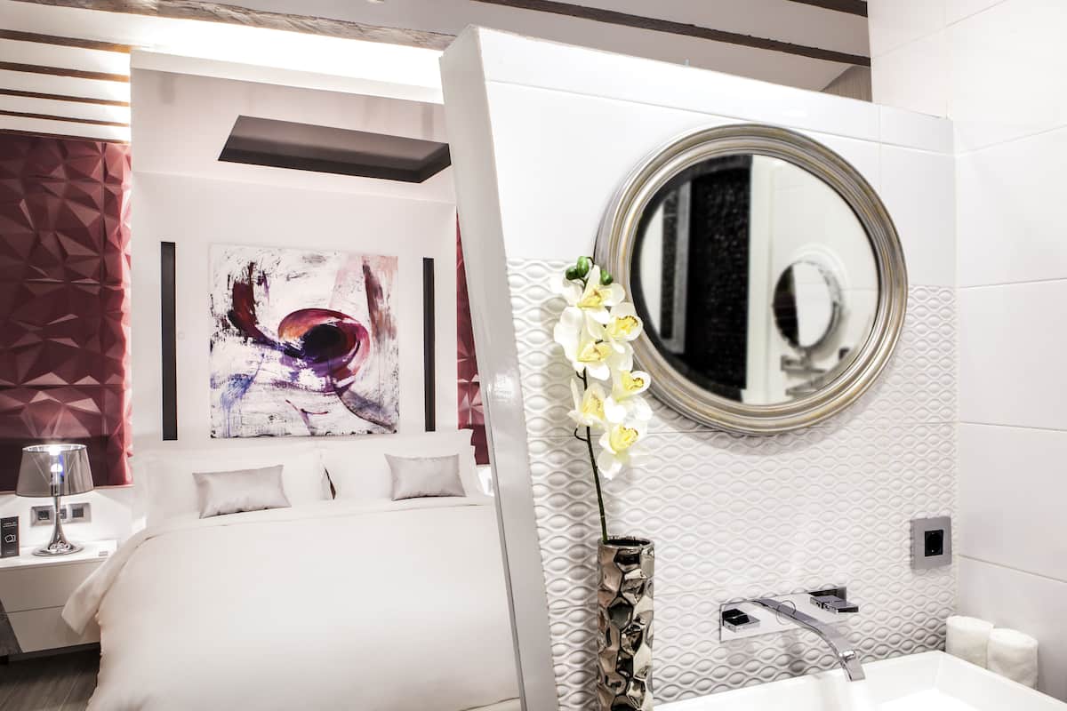 A modern hotel room with a bed, a round mirror, a vase with white flowers, and a white textured wall.