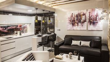 A modern living room with a black leather sofa, white armchairs, and a coffee table. There is a kitchen area with white cabinets and black marble countertops.