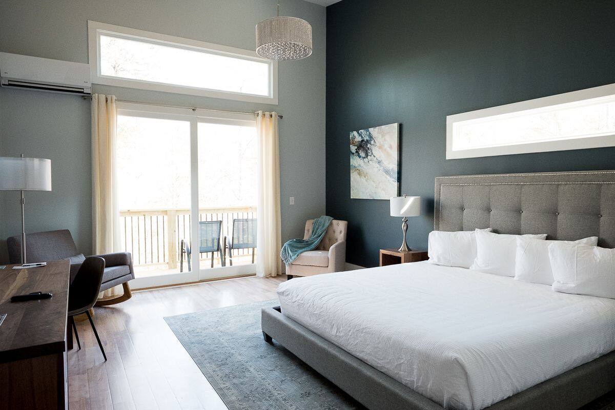A bedroom with a large bed, a desk, a chair, a lamp, a painting, and a view of the outdoors.
