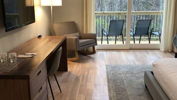 A modern hotel room with a wooden desk, a flat-screen TV, a comfortable chair, a bed, and a balcony with chairs.