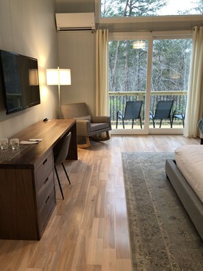 A modern hotel room with a wooden desk, a flat-screen TV, a comfortable chair, a bed, and a balcony with chairs.
