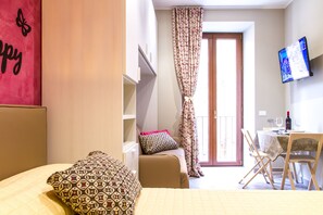Habitación de hotel con cama, una mesita con silla, televisor en la pared y balcón con cortinas.