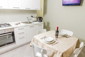 Una cocina compacta con armarios blancos, estufa a gas, hervidor, y una mesa de comedor con platos, una botella de vino y vasos.
