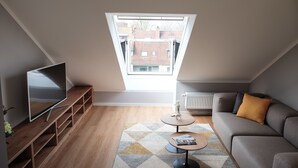 A modern living room with a sloped ceiling, a flat-screen TV on a wooden stand, a grey sofa with a mustard pillow, a small round table with a magazine, and a patterned rug. - Glück Auf Appartements Rüttenscheiderstraße (Essen)