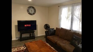 Een woonkamer met een televisie waarop 'NETFLIX' staat, een klok aan de muur, een bank, een ventilator en een kamerkleed met een patroon.