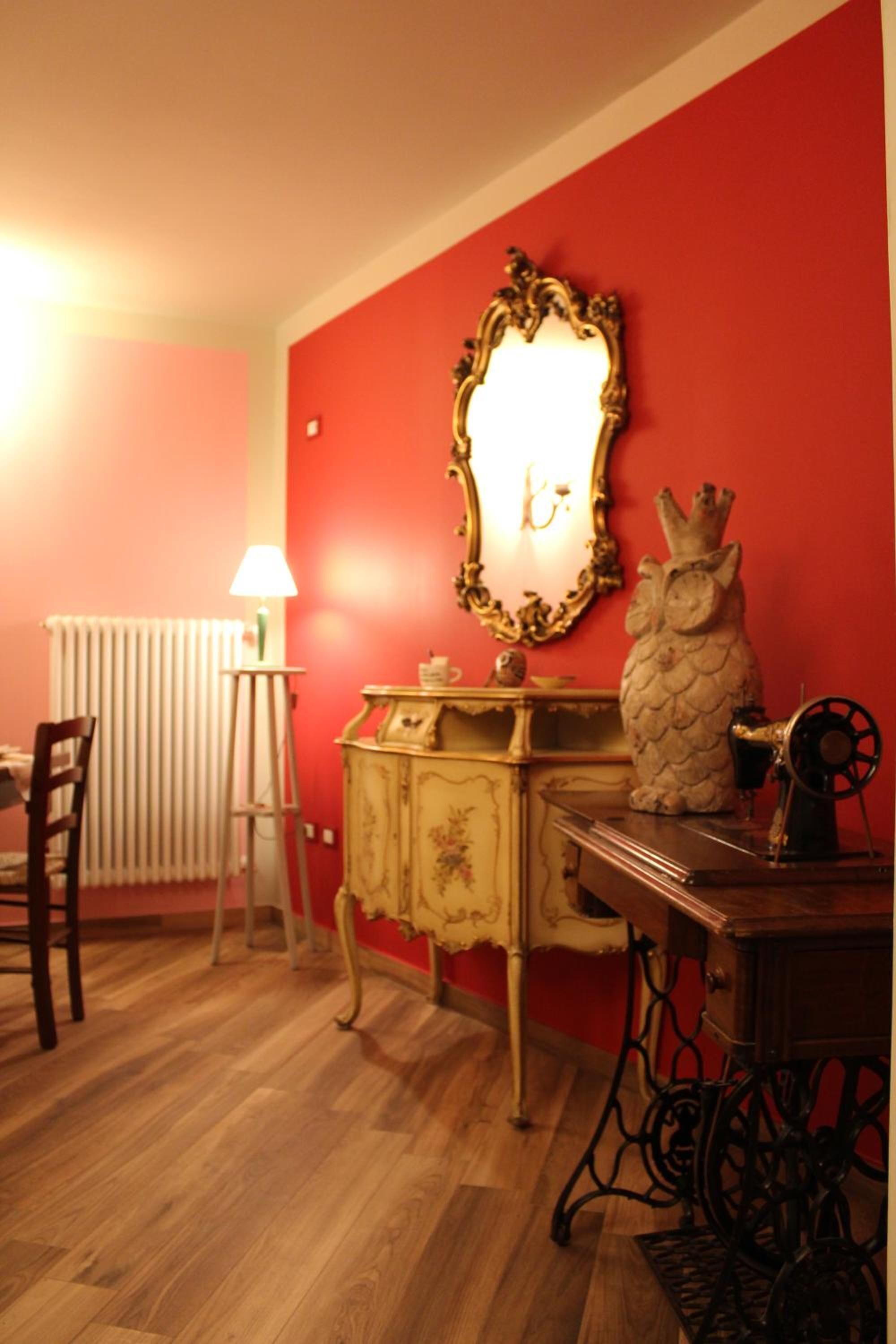 Um quarto com parede vermelha, assoalho de madeira, uma máquina de costura antiga, um abajur e um espelho.