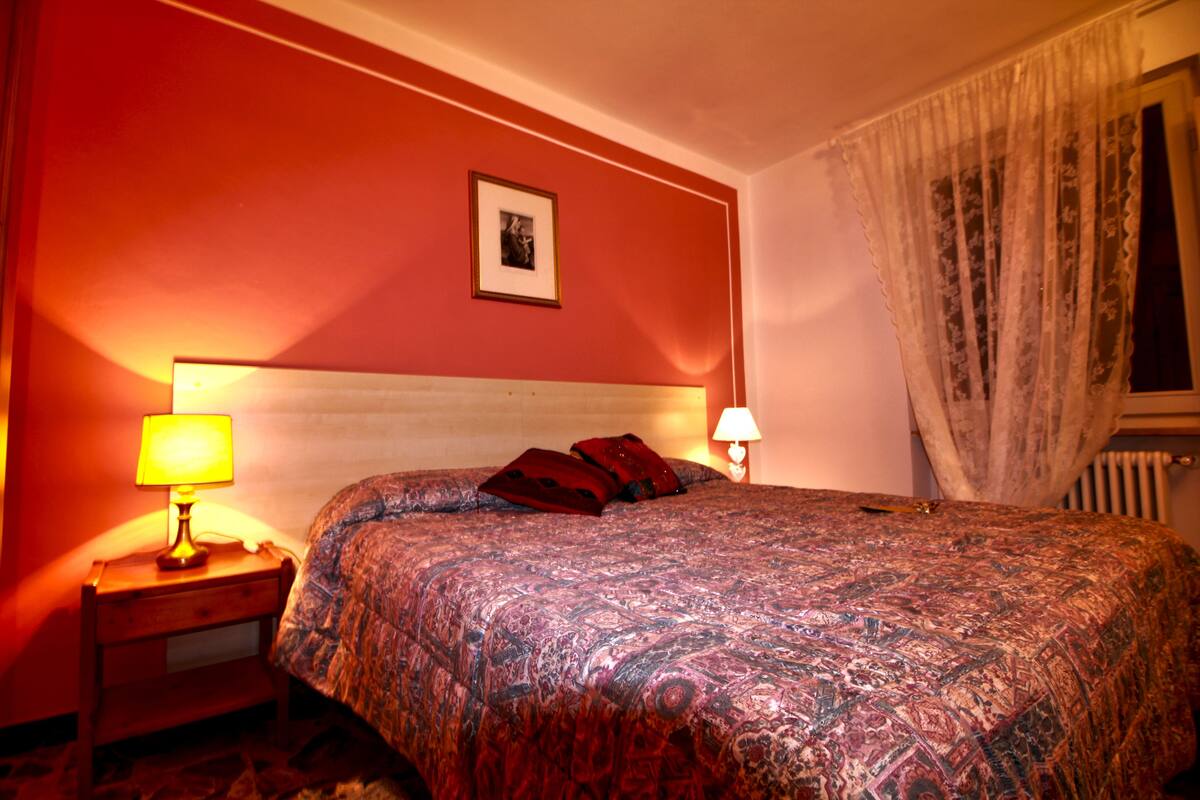 Ein Hotelzimmer mit einem großen Bett, zwei Nachttischlampen, einem gerahmten Bild an der Wand und einem Fenster mit durchsichtigen Vorhängen.