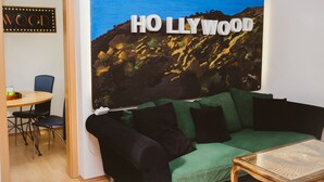 A living room with a green sofa, a small wooden coffee table, and a painting of a mountain landscape with the text "HO LLY WOOD". - Glück Auf Appartements See Straße (Essen)