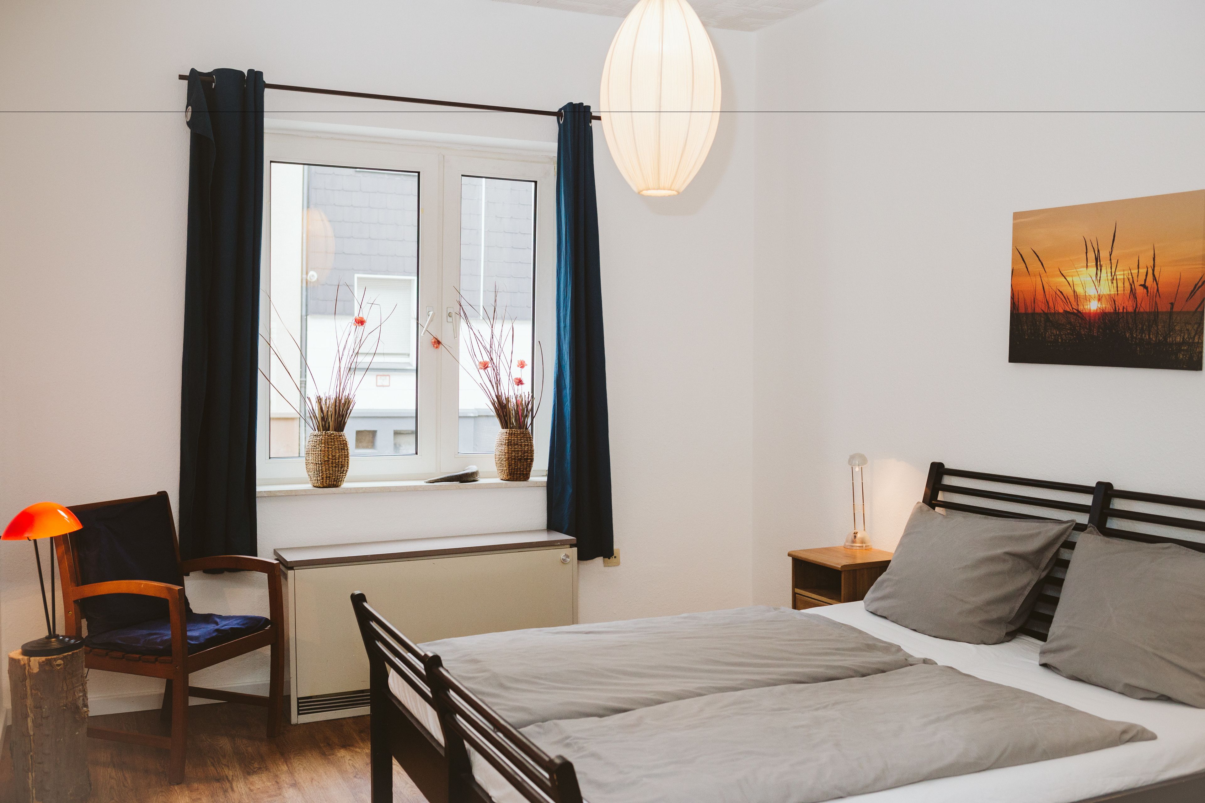 Une chambre à coucher comprenant un lit, une chaise, une table de chevet, une lampe, une fenêtre avec des rideaux, un vase de fleurs et une lampe fixée au mur.