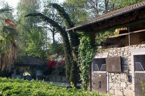 A stone building with a tiled roof, a covered outdoor seating area, and a lush garden with various plants and trees. - La Cucchetta (Dormelletto)