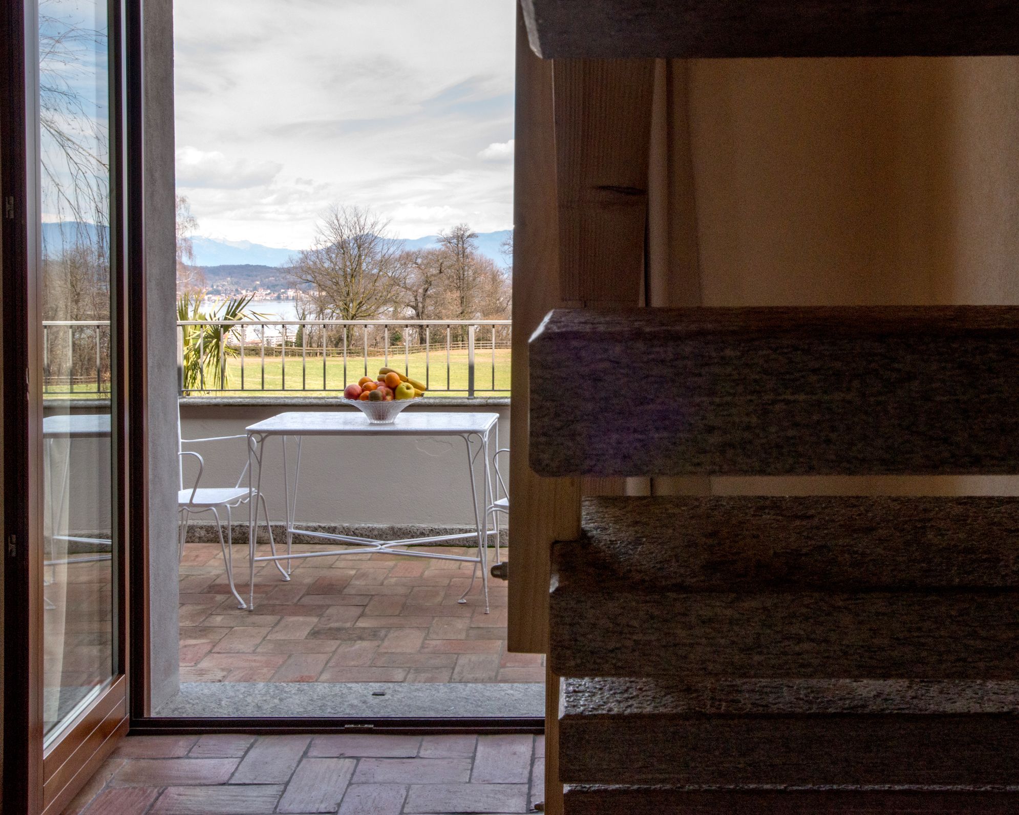 Vue depuis une pièce sur un espace extérieur comprenant une table et des chaises, un saladier de fruits et un garde-corps donnant sur un paysage avec des arbres et des montagnes.