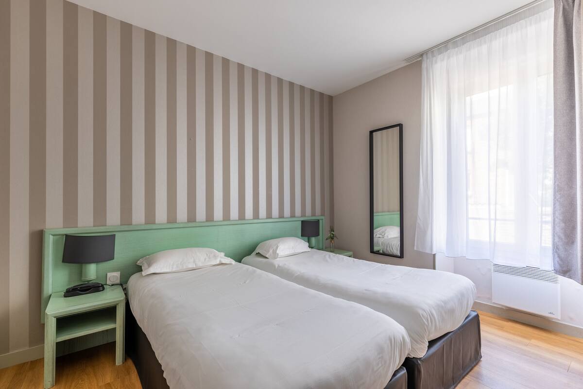 A hotel room with two beds, a nightstand with a lamp, a mirror, and a window with curtains.