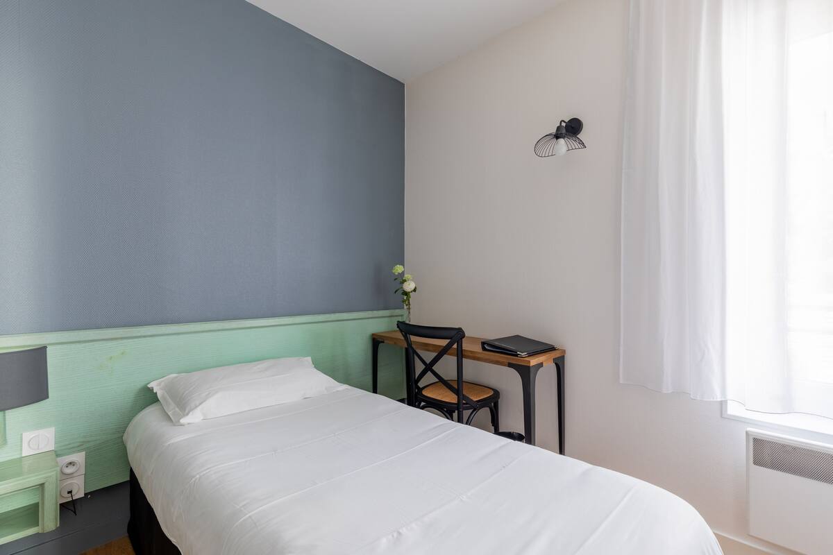 A bedroom with a bed, a desk with a chair, a small bedside table, and a wall-mounted lamp.