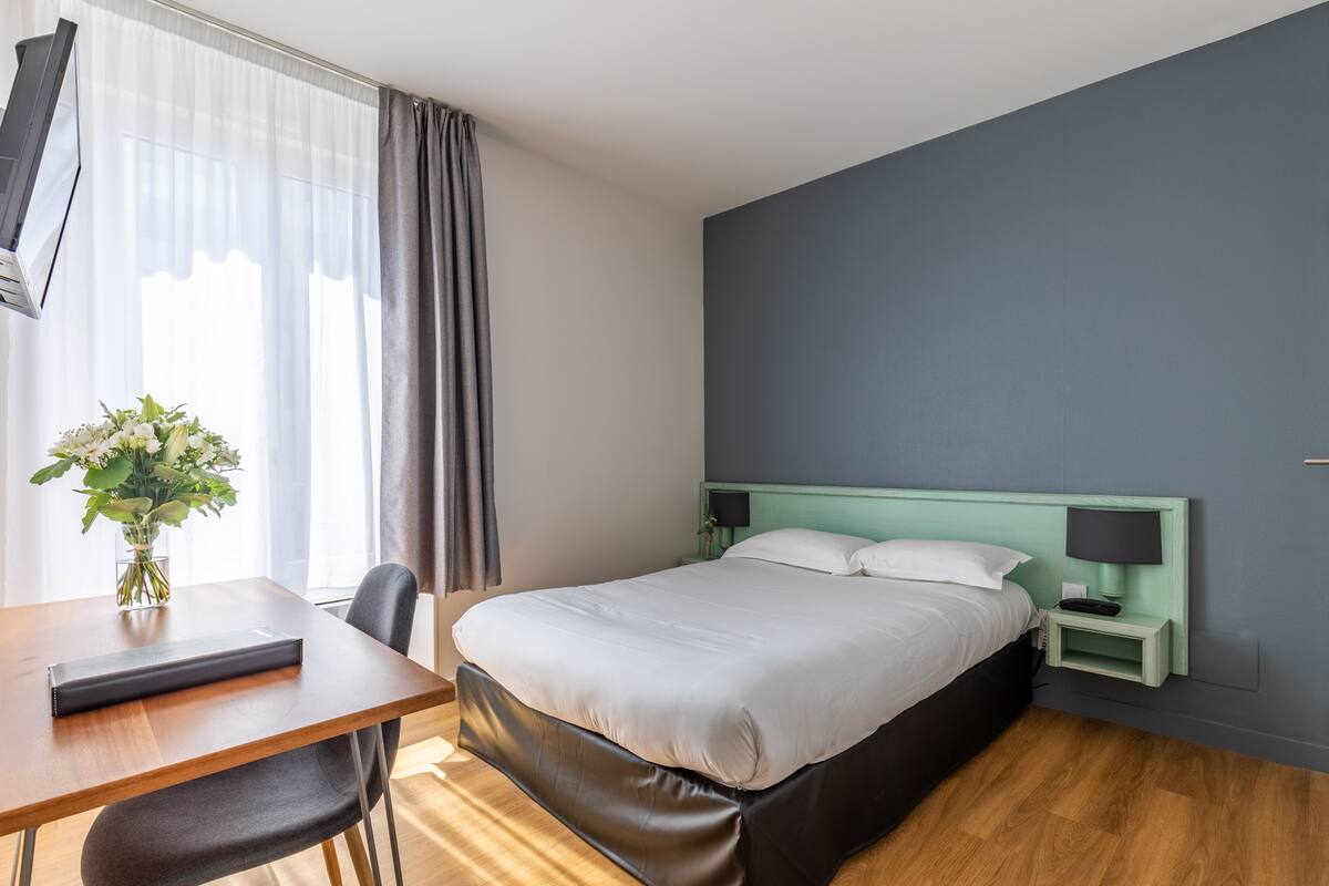 A hotel room with a bed, a desk, a chair, a nightstand, a lamp, and a window with curtains.