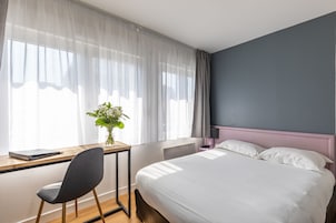 Une chambre d’hôtel avec un lit, un bureau, une chaise et une fenêtre avec des rideaux légers.