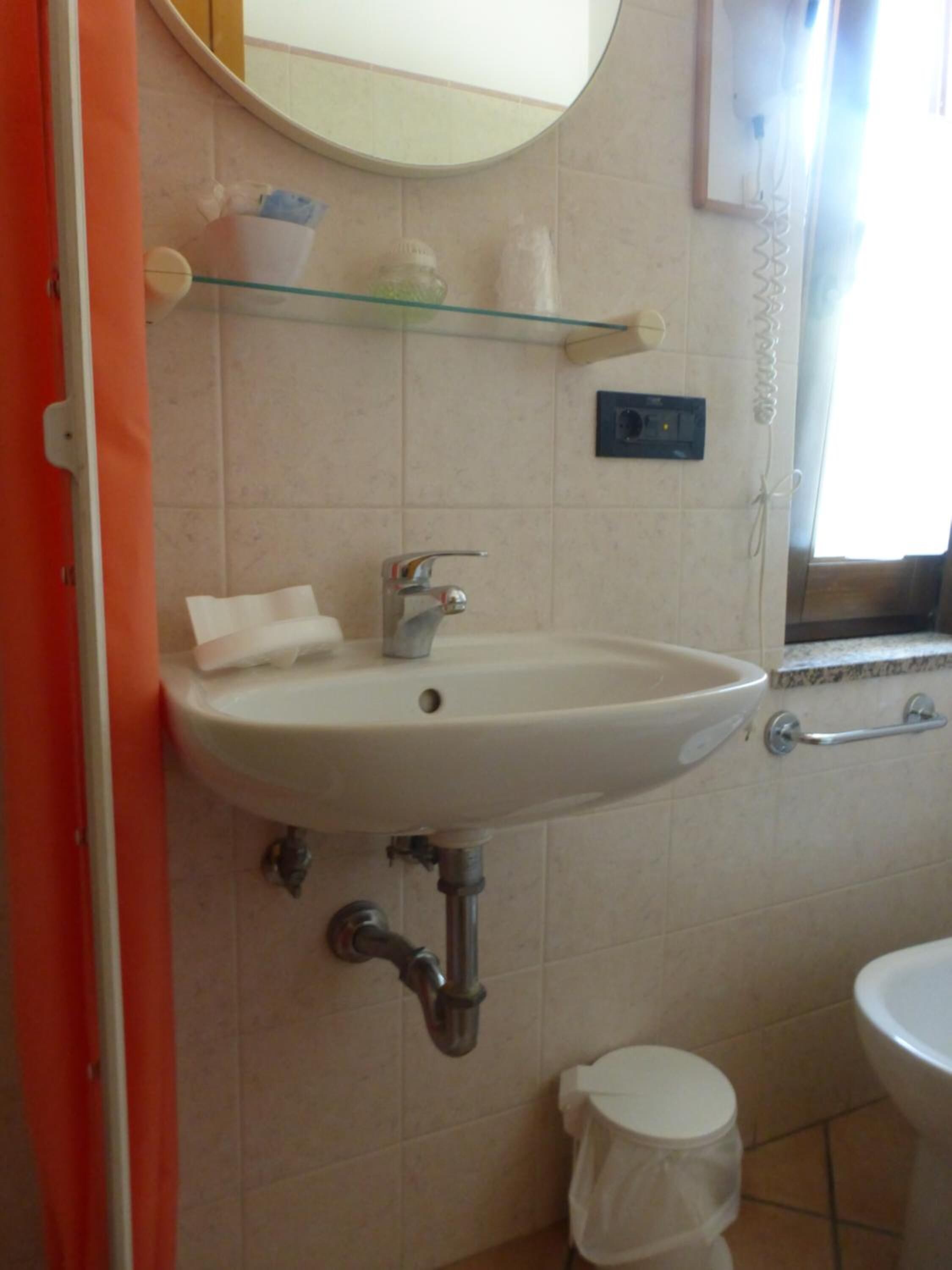 Un baño con lavamanos blanco, espejo y retrete. Hay un estante de vidrio sobre el lavamanos con algunos objetos.