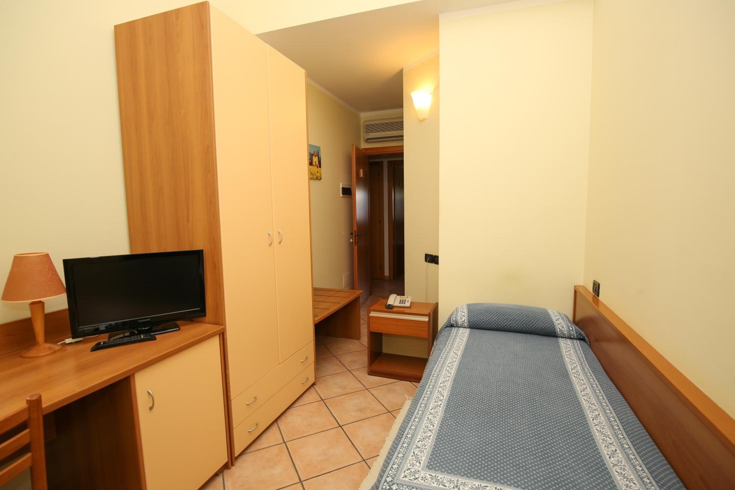 A small, neatly arranged bedroom with a bed, a desk with a television, a wardrobe, and a bedside table with a lamp.