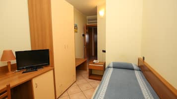 A small, neatly arranged bedroom with a bed, a desk with a television, a wardrobe, and a bedside table with a lamp.
