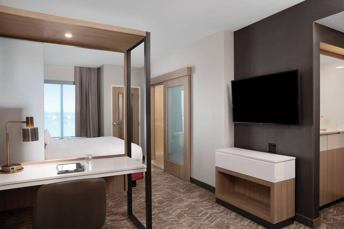 Habitación de hotel con cama, mesita de noche, lámpara, televisor y baño con lavamanos y espejo.