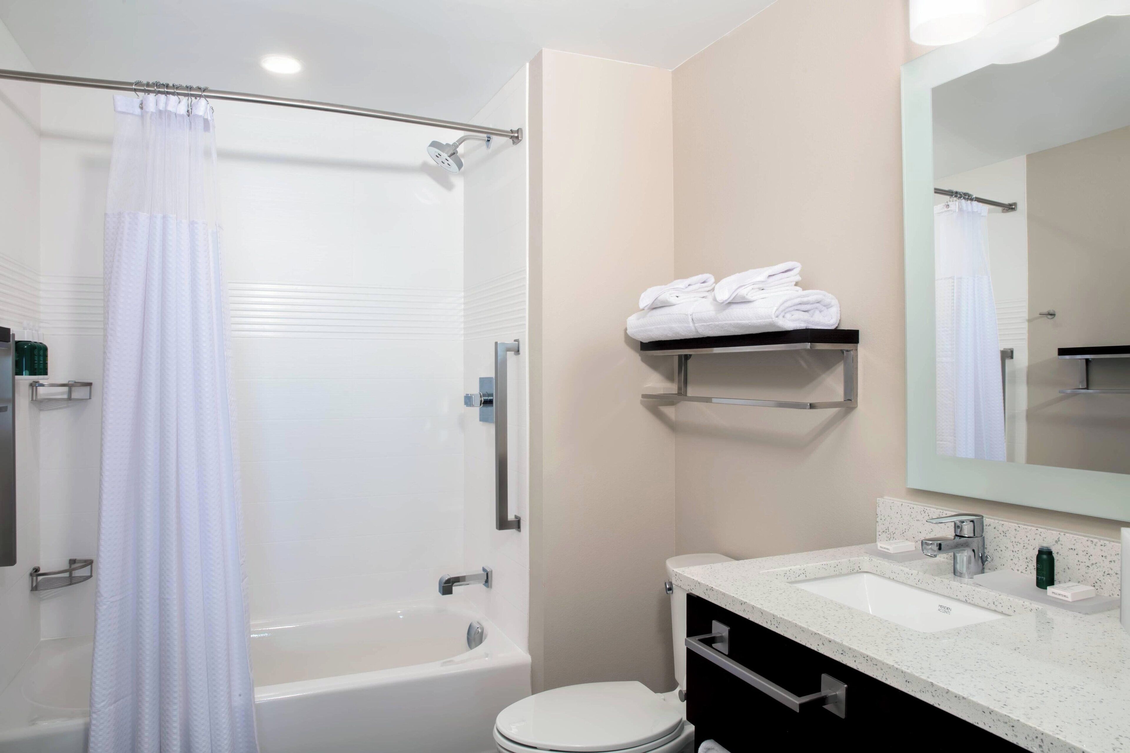 A modern bathroom with a bathtub, toilet, and sink. There is a shower curtain, a towel rack with towels, and a mirror above the sink.