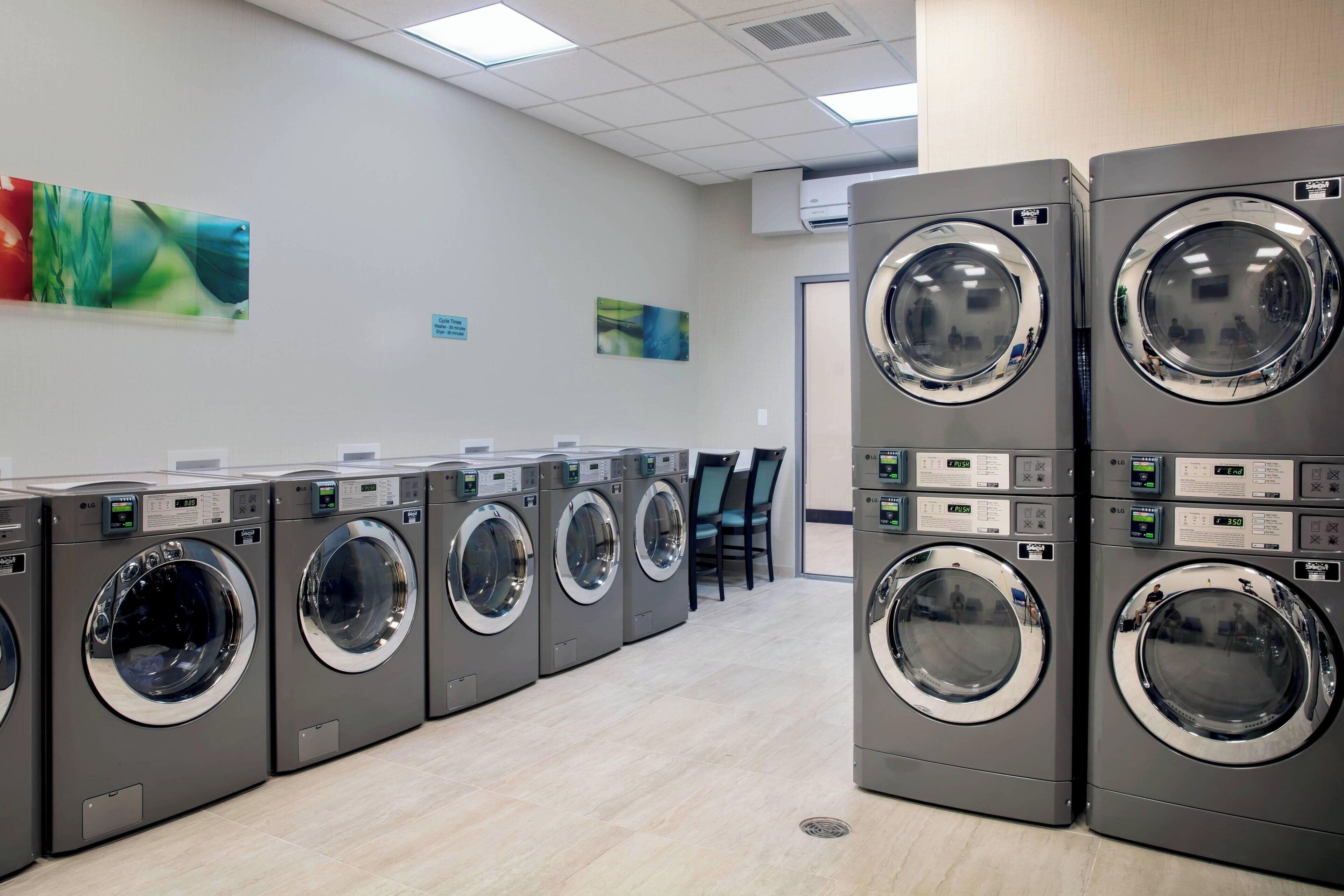 A row of modern, grey front-loading washers with digital displays, arranged in a clean, tiled-walled laundry room with chairs and artwork on the walls.