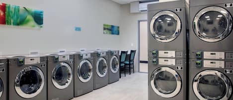A row of modern, grey front-loading washers with digital displays, arranged in a clean, tiled-walled laundry room with chairs and artwork on the walls.