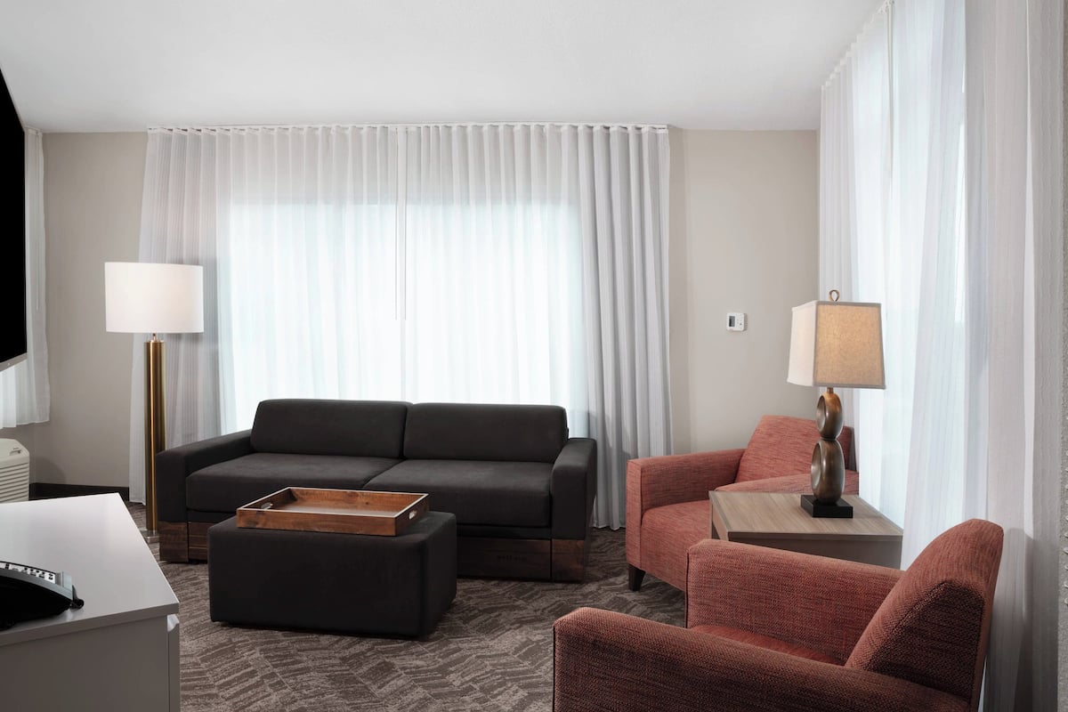 Una habitación de hotel moderna con un sofá gris oscuro, un sillón rojo, una mesita de madera con una lámpara y un ventanal con cortinas translúcidas.
