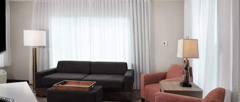 Una habitación de hotel moderna con un sofá gris oscuro, un sillón rojo, una mesita de madera con una lámpara y un ventanal con cortinas translúcidas.