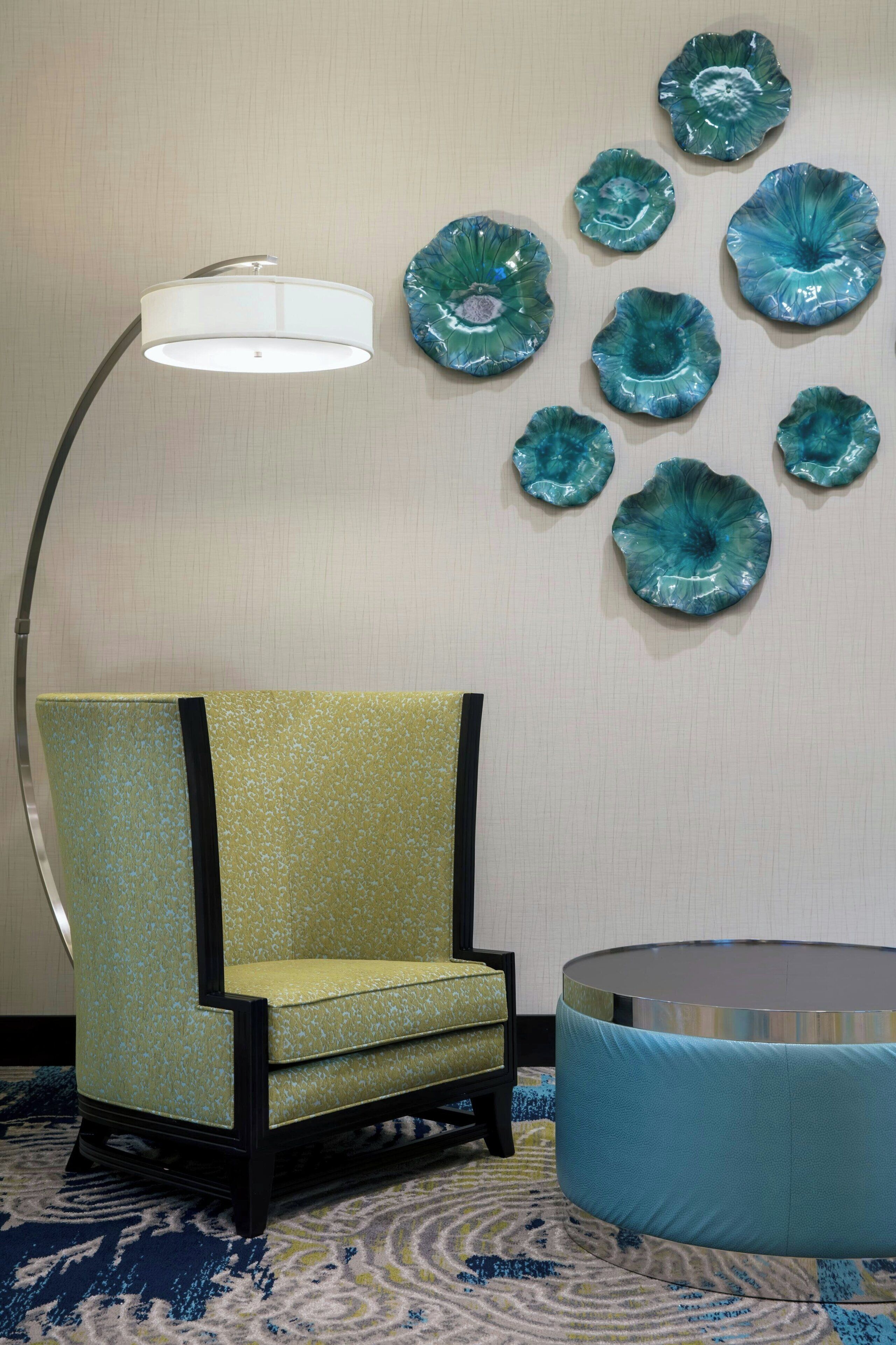 A room with a green upholstered armchair, a round blue ottoman, a floor lamp, and a wall decorated with turquoise glass art pieces.
