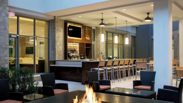 A modern indoor patio with a fire pit table, seating, a bar area, and large windows. - SpringHill Suites by Marriott Orlando Theme Parks/Lake Buena Vista (Orlando)