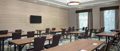 Un salón de conferencias con varias mesas y sillas, un televisor de pantalla plana montado en la pared y amplias ventanas con cortinas.