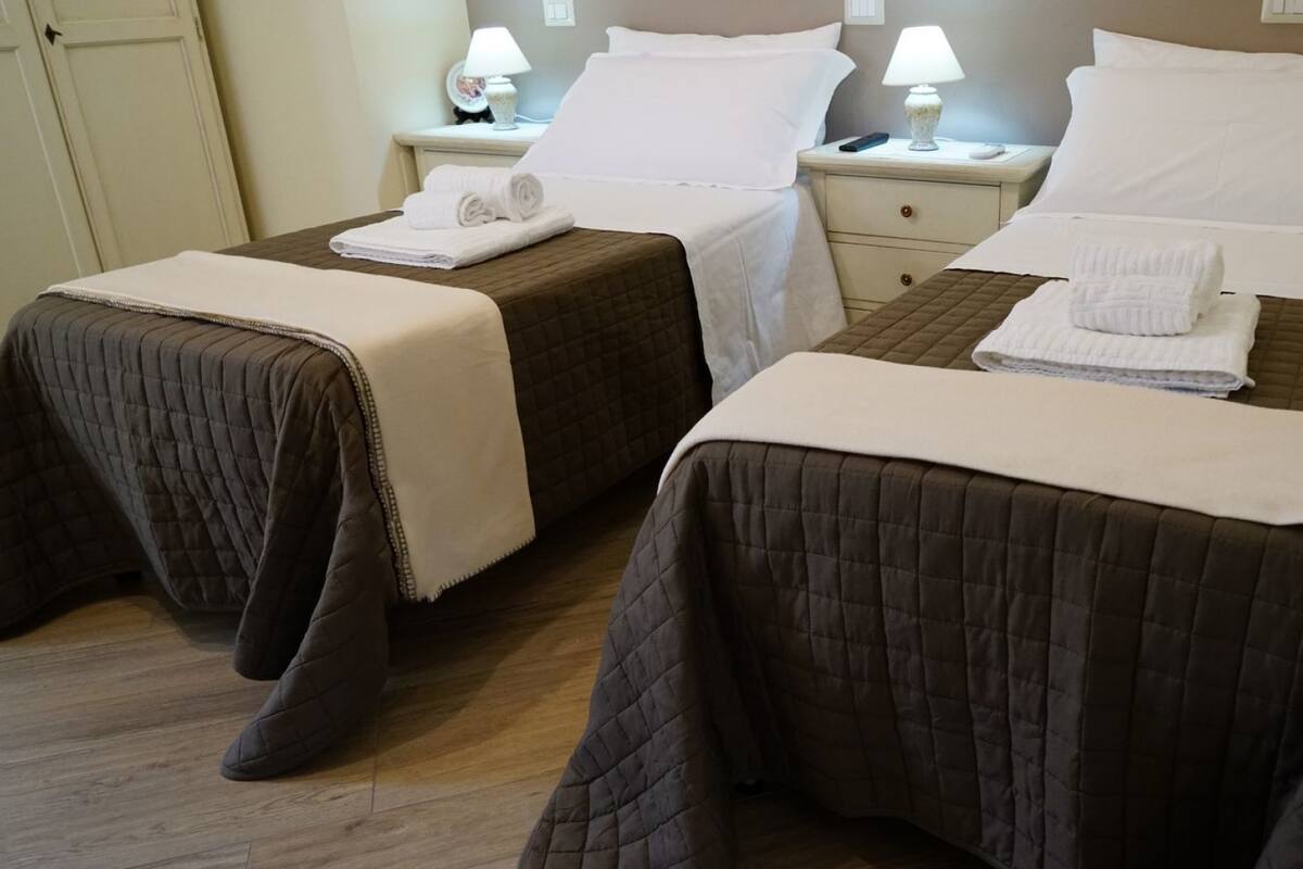 A hotel room with two beds, each with a quilted bedspread and white pillows. There are bedside tables with lamps and folded towels on each bed.