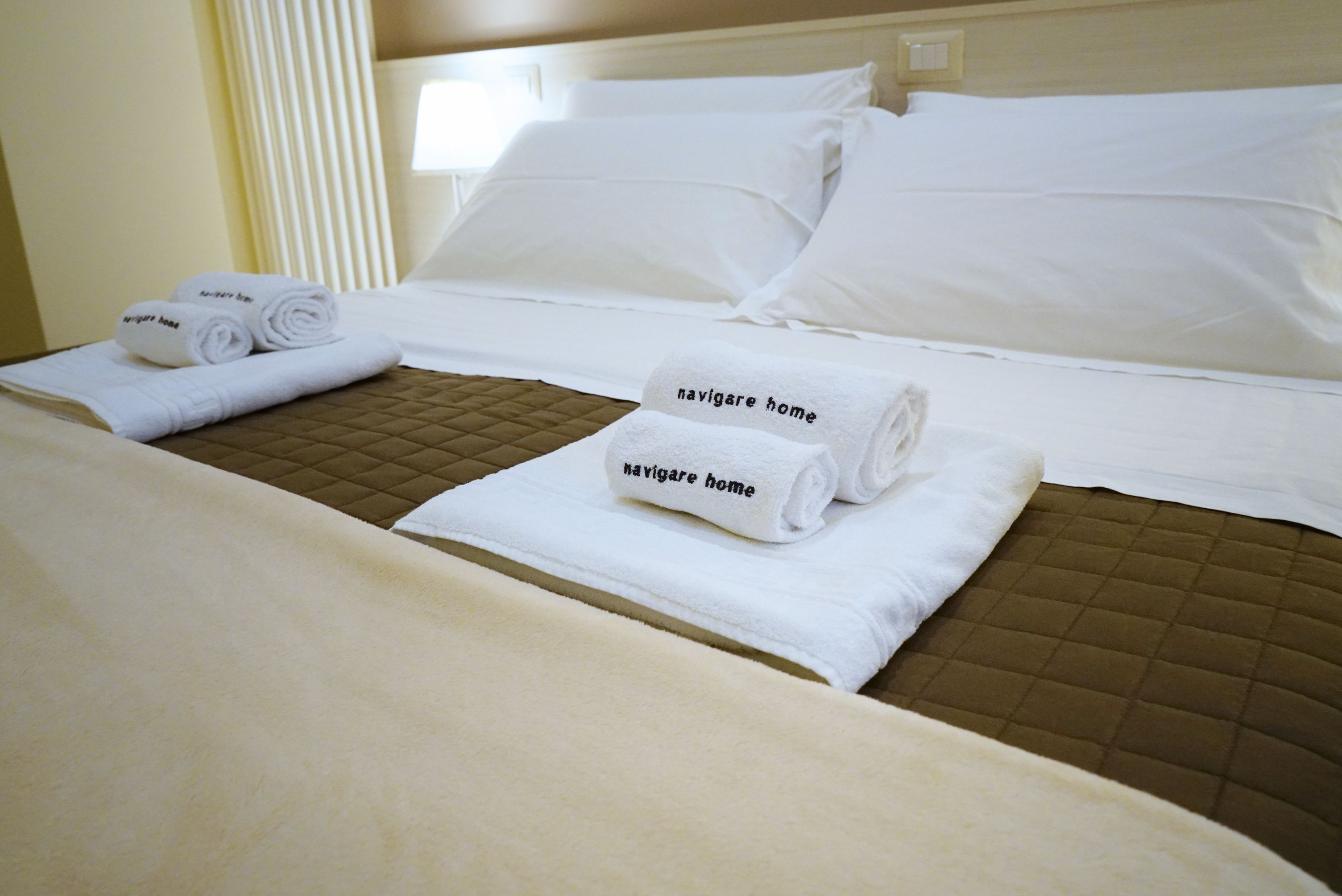 Habitación de hotel con una cama, dos almohadas y dos toallas enrolladas con el texto 'navigare home'.