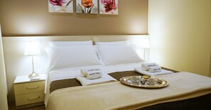 Quarto de hotel bem arrumado com uma cama grande, duas luminárias de cabeceira, um tabuleiro com itens de café da manhã e toalhas com o nome do hotel.