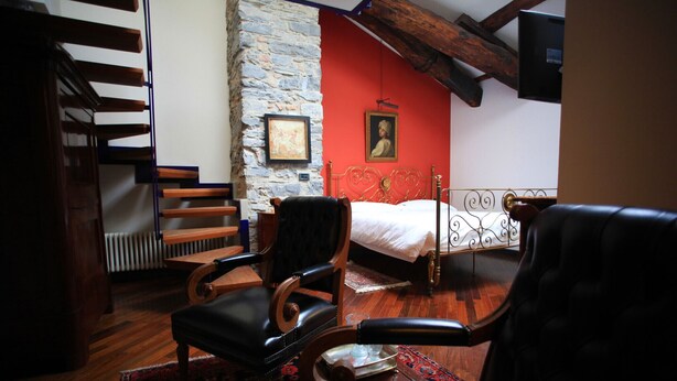 A room with a bed, two leather chairs, a wooden bench, a stone wall, and a red accent wall. - B&B Convento S. Antonio (Como)