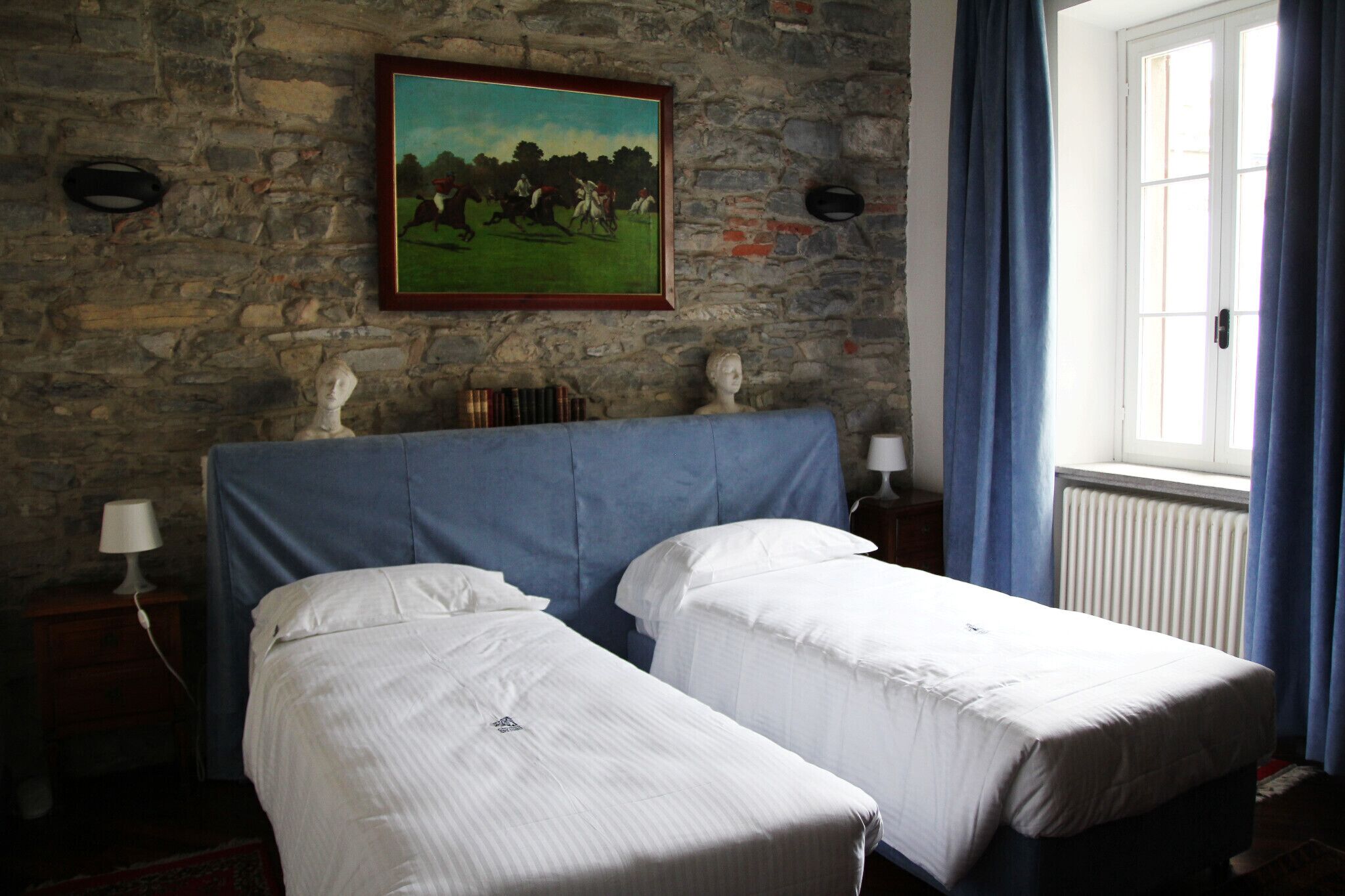 A room with two single beds, a stone wall, a painting, and a window with curtains.