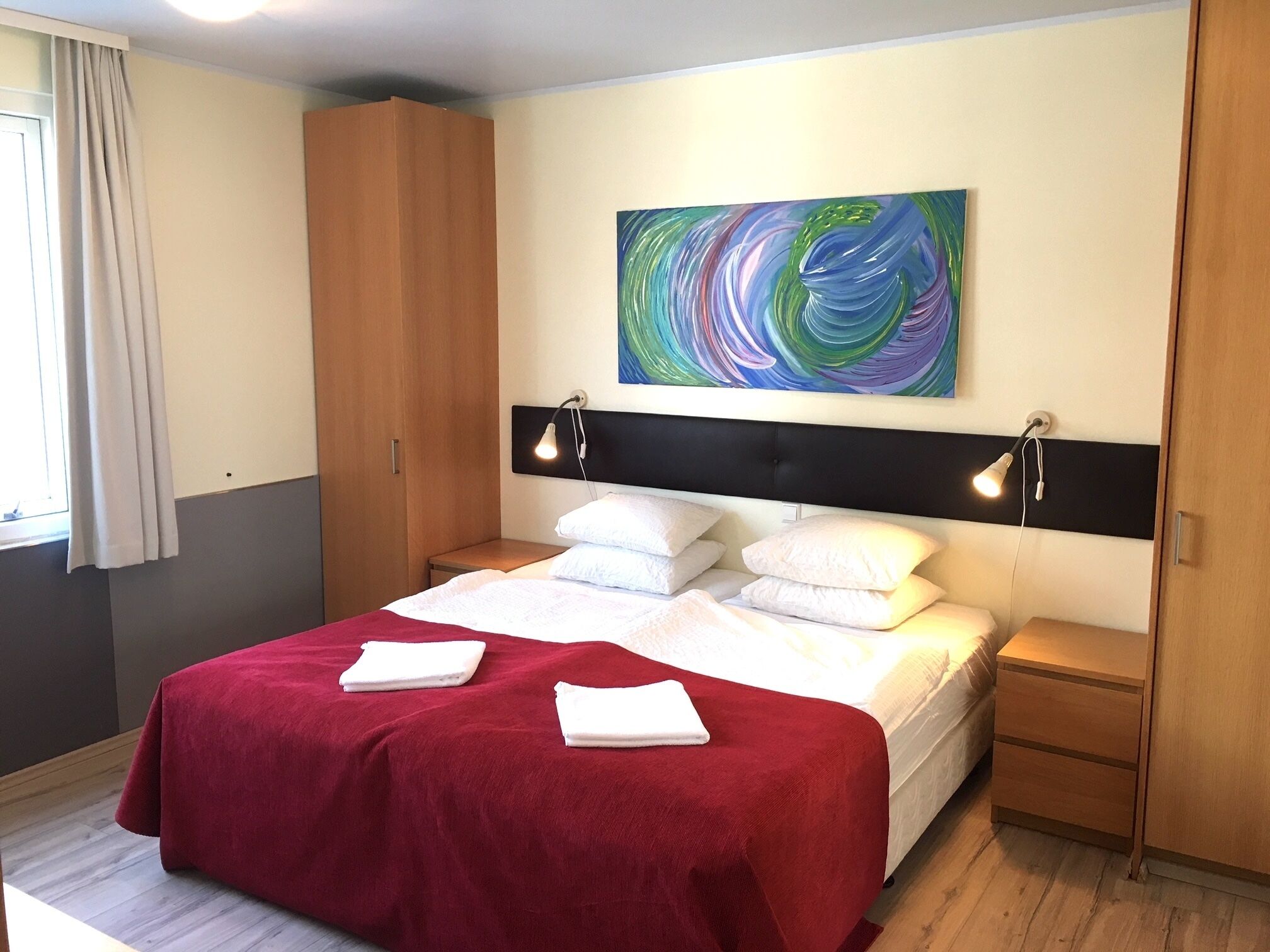 A neatly made bed with a red blanket and white pillows, a wooden wardrobe, a bedside table, and a colorful abstract painting on the wall.