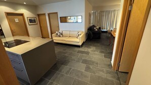 A modern kitchen with a central island, a living area with a sofa and a dining table, and a hallway with a door.