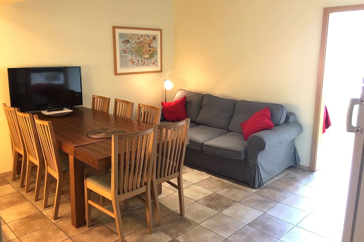 A dining area with a wooden table and chairs, a television, and a grey sofa with red cushions.