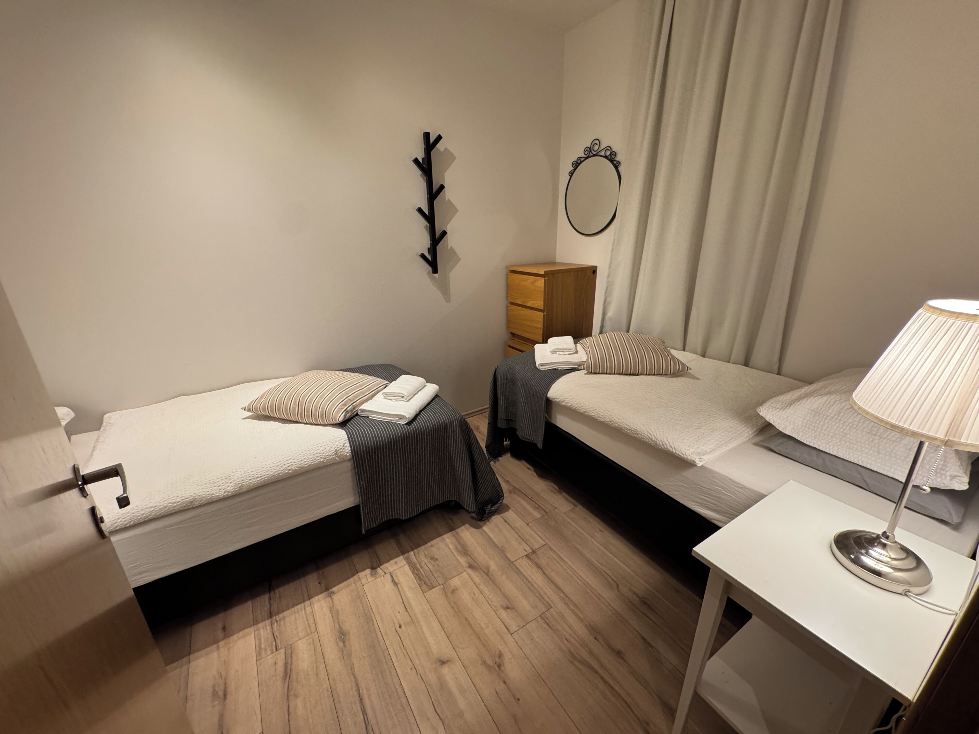 Quarto pequeno e bem iluminado com duas camas de solteiro, uma cômoda de madeira, mesa de cabeceira com abajur e um espelho na parede.