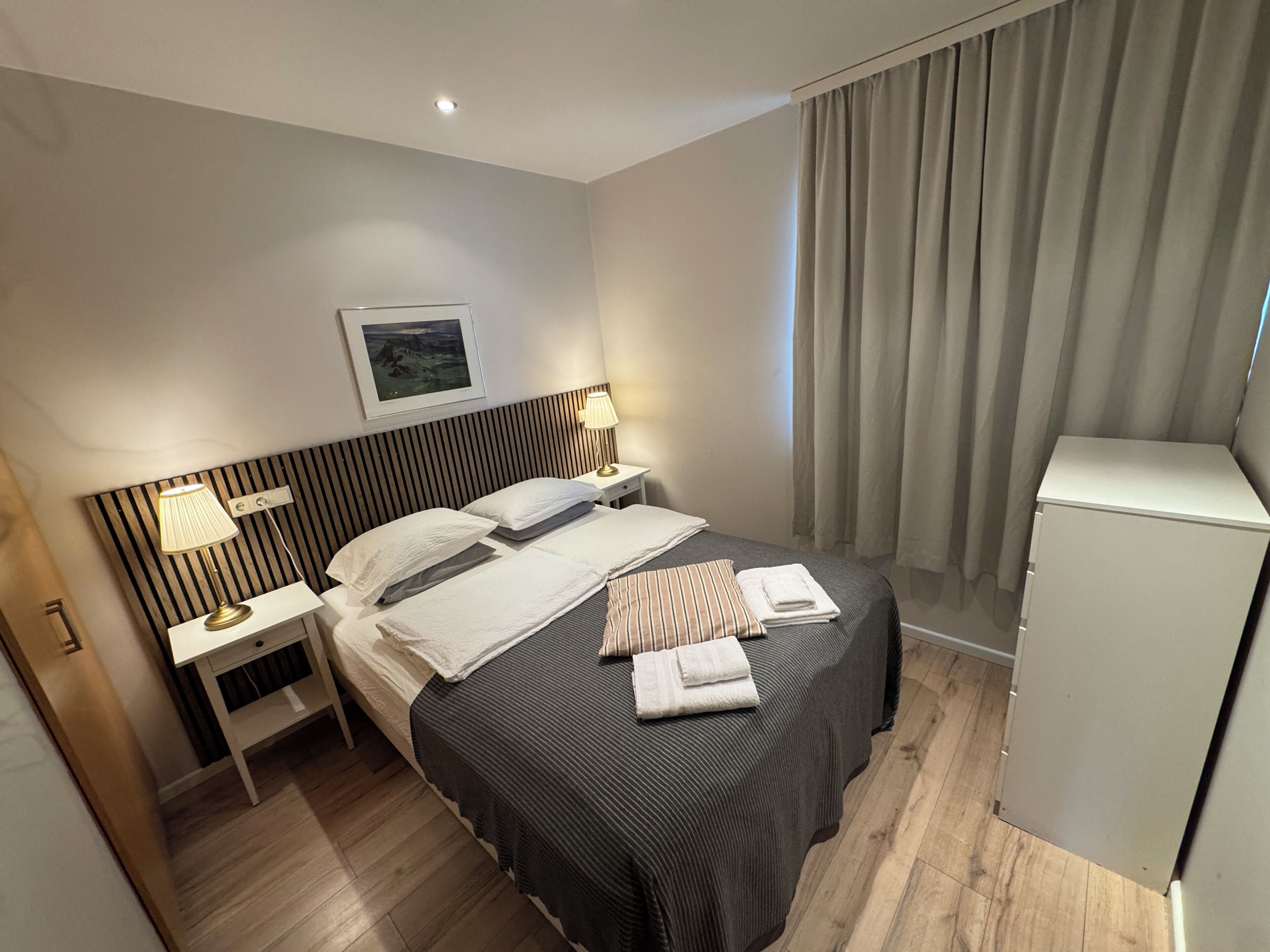 Een hotelkamer met een bed, bedlampen, een nachtkastje, een klein kastje en een raam met gordijnen.